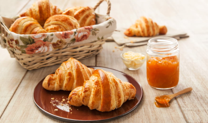 Croissant sobre um prato, acompanhados de um pote de geléia e com uma cesta de croissants atrás.